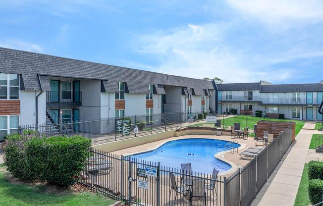 View of an apartment complex featuring an outdoor swimming pool surrounded by a fenced area. The pool is equipped with lounge chairs and tables. Two buildings with multiple units are visible in the background, set against a clear blue sky. Green landscaping surrounds the area, enhancing the inviting atmosphere.