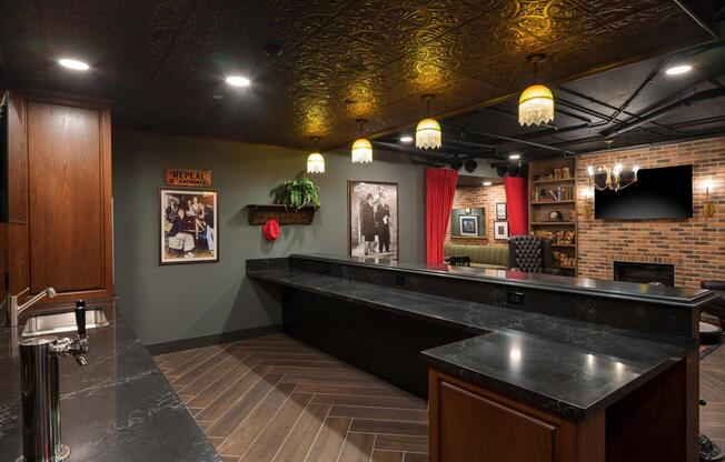 A stylish bar area featuring a dark countertop, vintage decor, framed black-and-white photos, and warm pendant lighting. The space includes a sink, wooden cabinetry, and a backdrop of red curtains and exposed brick, creating a cozy and inviting atmosphere for social gatherings.