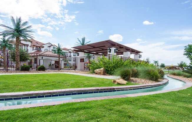A scenic outdoor area here at Andalucia Villas with lush green lawn, landscaped plants, and a winding water feature, complemented by shaded pergola seating and Spanish-style buildings that create a peaceful, resort-like setting.