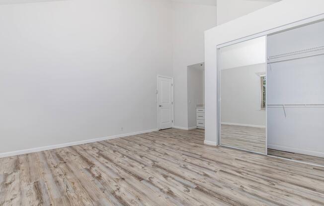 A spacious, empty room with high ceilings and a light-colored wall. Features include a wooden floor, a large mirrored sliding closet door, and a doorway leading to another area. The space is bright and minimalist, suitable for various uses.