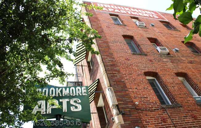 Upshot of large brick building in Pasadena CA. Two large signs saying "Brookmore apts" and "Brookmore Apartments" on building with green trees in front.