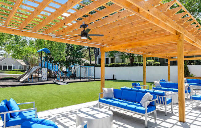 A wooden pergola with a ceiling fan provides shade for a patio with blue chairs and tables.
