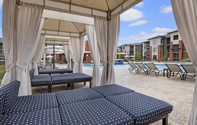 Poolside cabana here at 49 West Apartments with polka-dotted lounge chairs, surrounded by white curtains, overlooking a pool and modern apartment buildings under a blue sky.