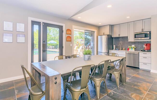 A modern kitchen and dining area featuring a large rectangular table with metal chairs, stainless steel appliances, and sleek cabinetry. There are sliding glass doors leading to an outdoor space with a playground visible. The walls have framed documents and the flooring is composed of dark slate tiles.