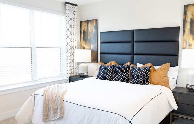 A bed with a white comforter and a white blanket is in the foreground of a room with a black headboard and a window with white blinds.