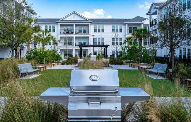 A silver grill is in the foreground of a grassy area in front of a white building.