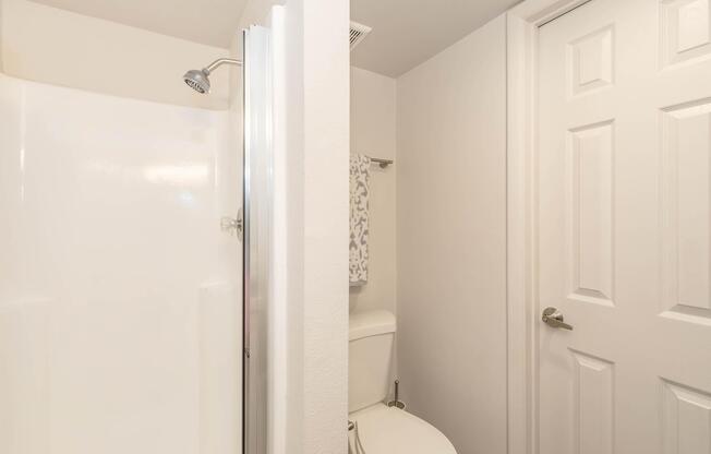 A bathroom featuring a shower on the left side and a toilet on the right. The shower has a clear glass door, and there is a towel hanging nearby. The walls are painted white, and there is a closed door leading out of the bathroom. The overall design is clean and modern.