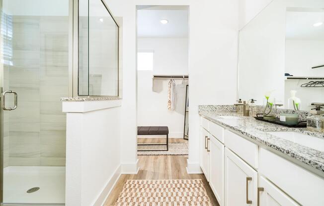 A bright primary bath features dual vanities, stone countertops, and a glass-enclosed shower, with a seamless connection to the spacious closet beyond.