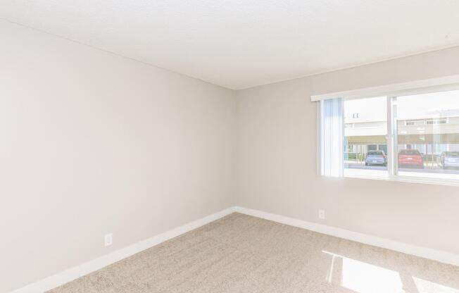 Empty room with light beige walls and carpet. A large window with white blinds lets in natural light, showing a view of parked cars outside. The space appears clean and well-maintained, suitable for moving in or decorating.