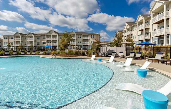 A large swimming pool with blue water and white lounge chairs in front of apartment buildings.