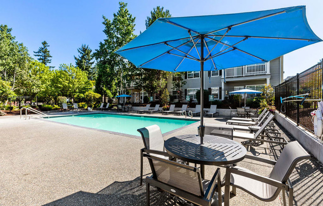 A blue umbrella shades a table and chairs by a pool.