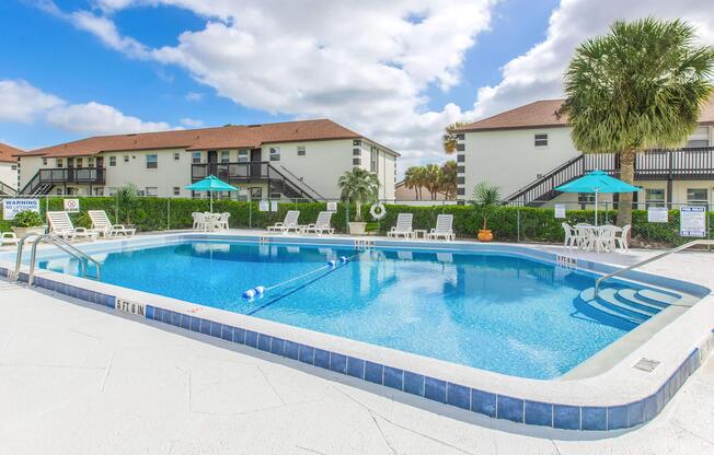 A clear swimming pool surrounded by lounge chairs and umbrellas, located in a residential area. Two-story apartment buildings are visible in the background, with green landscaping and palm trees. The sky is partly cloudy, creating a bright and inviting atmosphere.