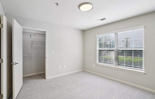 A room with a carpeted floor, a window with blinds, and a door leading to another room.