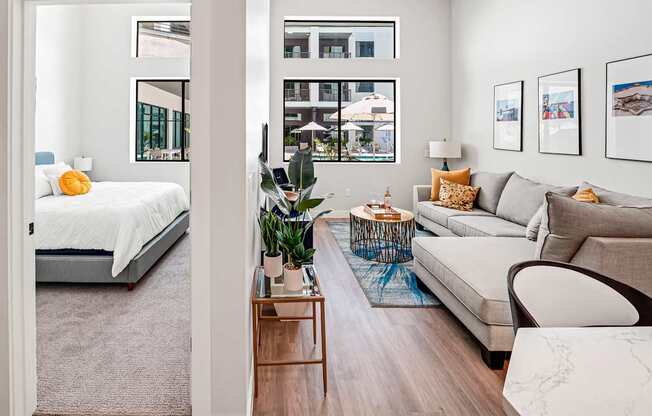 A modern living room with a grey sofa, a bed with white bedding, a wooden coffee table, and a large window with a view of the city.
