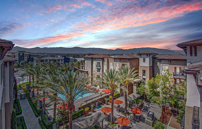 a courtyard with tables and umbrellas and a pool with a sunset in the background