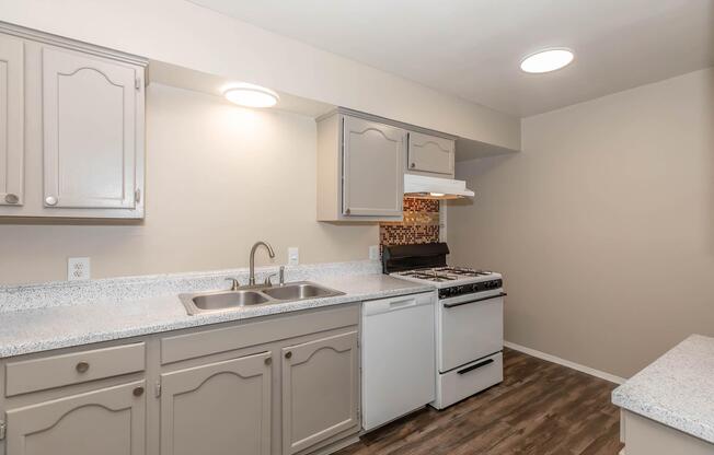 A modern kitchen with light gray cabinets, a white gas stove, and a double sink. The counter has a speckled finish, and there is a patterned backsplash behind the stove. The walls are painted a neutral color, and the lighting is bright and even, creating an inviting space.