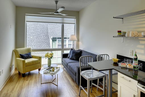 Living area with grey couch and yellow accent chair, large window and ceiling fan. Glimpse of breakfast nook and open kitchen shelves.