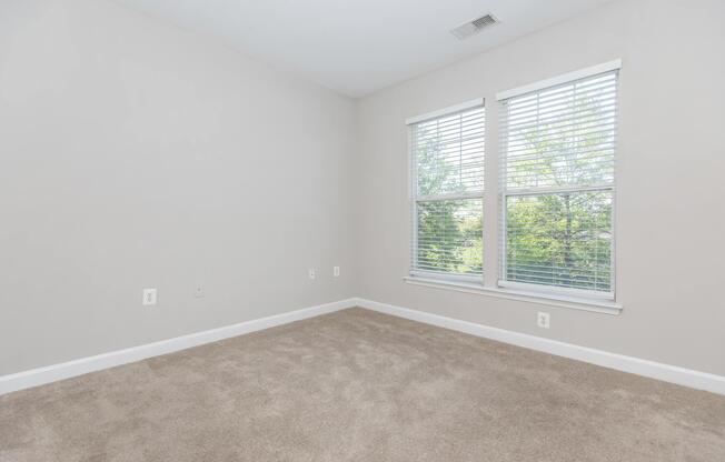 Empty room with light gray walls and beige carpet. Two windows with white blinds allow natural light to enter, revealing a view of greenery outside. The room is spacious with no furniture, providing a clean and minimalist aesthetic.