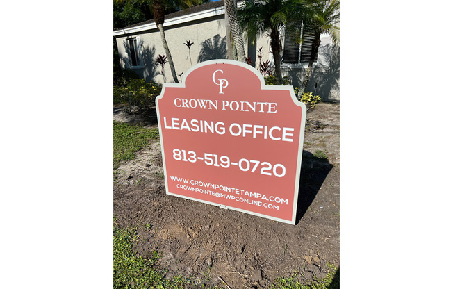 Sign for Crown Pointe Leasing Office displaying the phone number 813-519-0720 and website crownpointetampa.com. The sign is positioned in front of a building with greenery surrounding it.