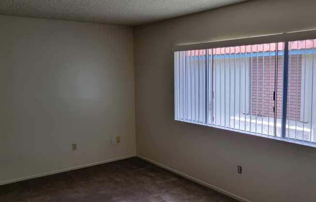 Carpeted bedroom with large window and vertical blinds at Magnolia Apartments in Riverside, California.