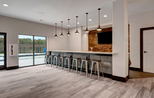 A modern bar area here at The Park featuring a long counter with metal barstools, warm pendant lighting, wood accent wall, and large windows that create a bright, inviting social space.
