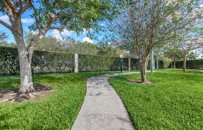 A curved concrete pathway lined with green grass and trees on either side, leading through a park-like area. The background features a decorative black fence and bright blue sky with a few clouds. Sunlight casts shadows on the pathway, creating a tranquil outdoor setting.