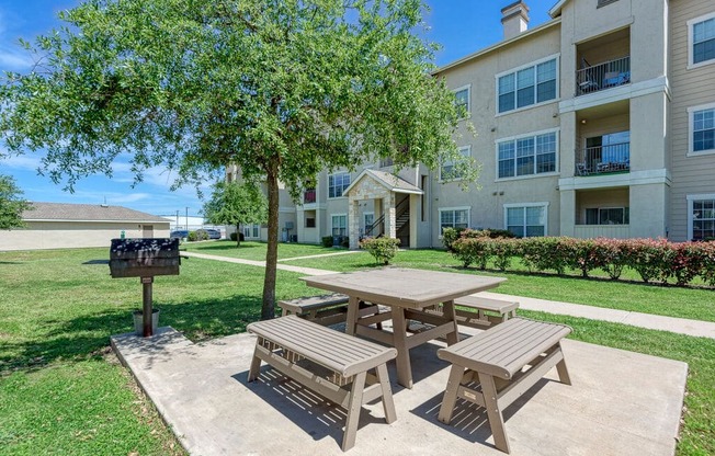 A wooden picnic table is in the foreground of a grassy area with apartment buildings in the background.