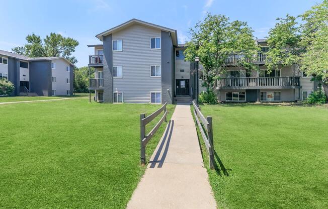 A view of a residential apartment complex with two buildings surrounded by green grass. A concrete pathway leads to the entrance, flanked by wooden fences and trees. The sky is clear and blue, creating a welcoming atmosphere.