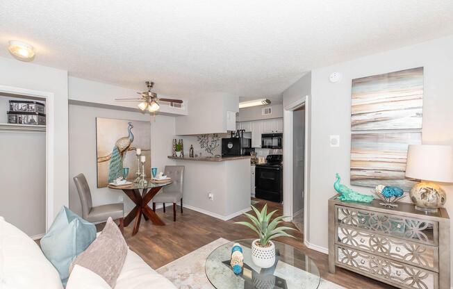 A modern living space featuring a cozy seating area with a white sofa and decorative pillows, a dining table set for two, and a kitchenette. The walls are painted in light grey, and there are artworks with a peacock theme. Natural light illuminates the room, complemented by stylish decor and a potted plant.
