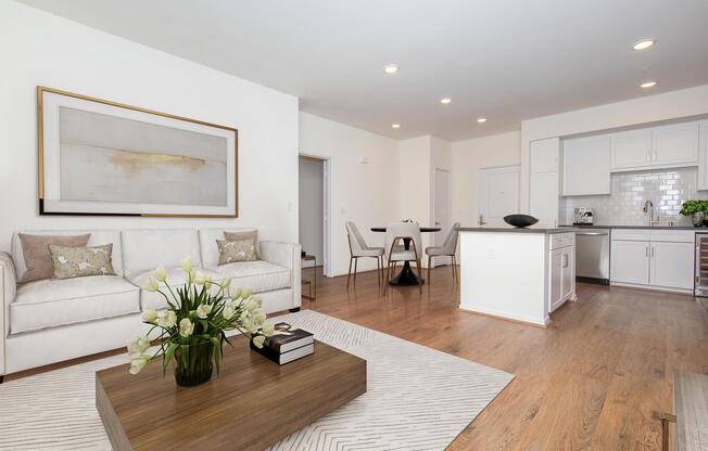 A modern living room and kitchen area featuring a white sofa with decorative cushions, a coffee table with books and a vase of flowers, hardwood flooring, and a well-lit open layout. The kitchen has white cabinets and appliances with a stylish backsplash. A dining area with a round table and chairs is visible in the background.