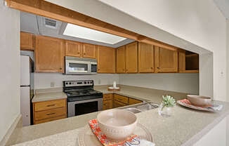 A cozy, functional kitchen here at Summertree Place featuring warm wood cabinetry, spacious countertops with a breakfast bar opening to the living area, a stainless finish appliance set, a double-basin sink, and simple place settings that highlight the practical layout and everyday convenience of the space.