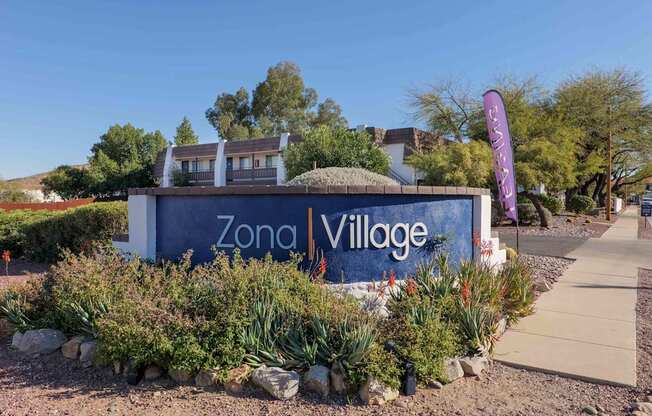 A sign for Zona Village is surrounded by plants and rocks.