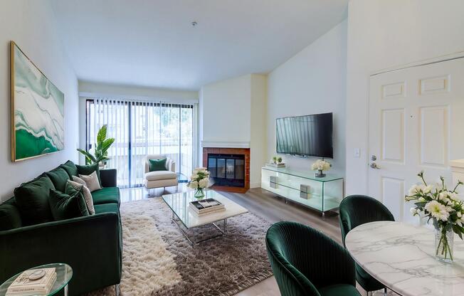 A modern living room featuring a green sofa set, a glass coffee table on a soft rug, and a contemporary TV mounted on the wall. There are potted plants, a fireplace, and large windows with sheer curtains, allowing natural light to brighten the space. Elegant decor elements add to the stylish ambiance.