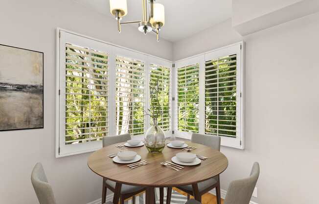 a dining area with a wooden table and chairs and multiple windows with white shutters