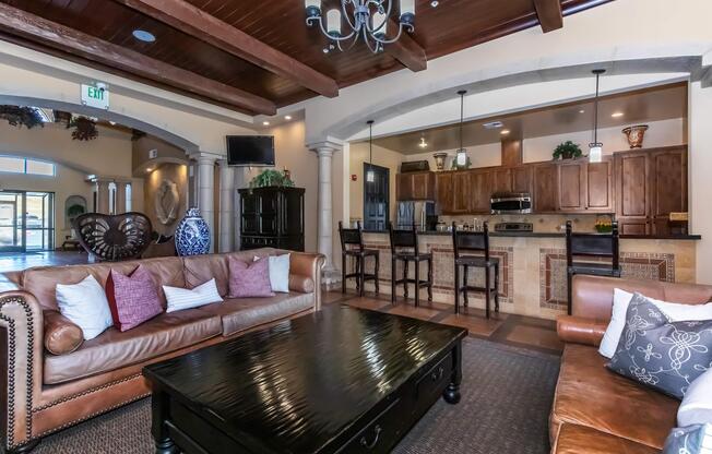 A spacious and elegantly decorated living area featuring a large coffee table, comfortable sofas with decorative pillows, and a stylish chandelier. The open kitchen in the background showcases wooden cabinets and modern appliances. Warm lighting and rich textures create a welcoming atmosphere.