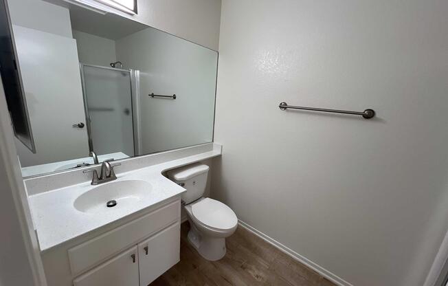 A clean, modern bathroom featuring a light-colored vanity with a sink, a toilet, and a large mirror. The walls are painted white, and there is a towel bar on one side. A glass shower is visible in the background, with wooden-style flooring throughout the space. Natural light enters from a window above the mirror.