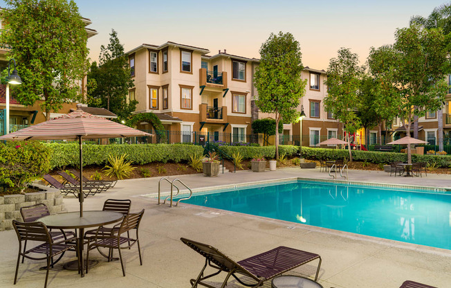 A pool surrounded by chairs and umbrellas in front of apartment buildings.