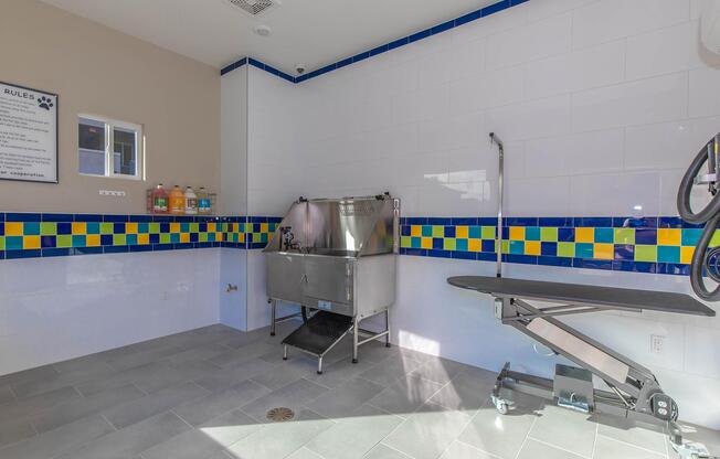 A clean, modern pet grooming area featuring a stainless steel grooming tub, a grooming table, and colorful tile accents on the walls. Shelves hold various pet care products, and the space is well-lit with white tiles and gray flooring, designed for convenience and efficiency in pet grooming.