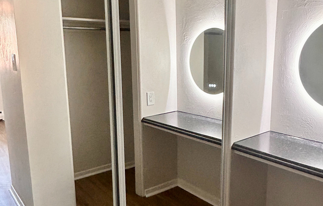 Modern apartment interior featuring a spacious walk-in closet with sliding mirrored doors and a stylish vanity area with illuminated round mirrors.
