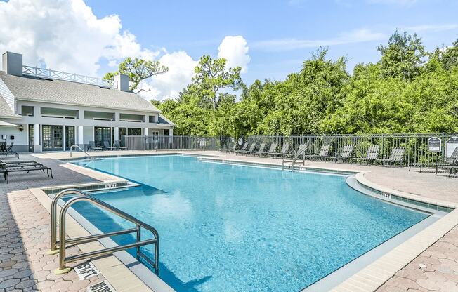 Swimming pool with large tanning deck, table with umbrella, lounge chairs, clubhouse in background surrounded by native landscaping