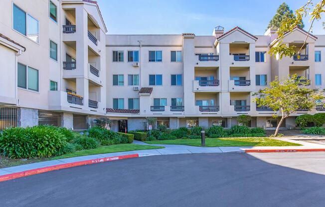 A residential apartment complex featuring a light-colored exterior with multiple balconies, surrounded by well-maintained landscaping. The foreground includes a paved parking area with a red curb. Clear blue skies are visible above, highlighting the sunny ambiance of the location.