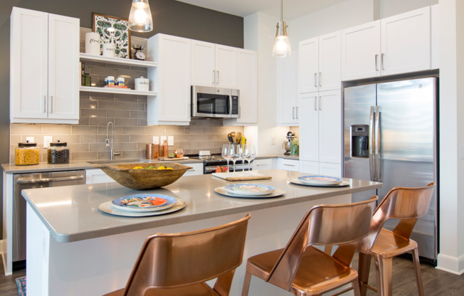 A bright kitchen with stainless steel appliances, white cabinets, built-in shelves, dark wood-style floors, and an island with three barstools.