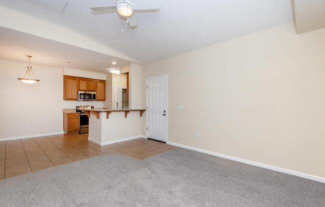 Spacious living area with light beige walls and plush gray carpet. A ceiling fan hangs overhead. To the left, a kitchen features wooden cabinets and stainless steel appliances. A doorway leads to a white door, and the room has natural light contributing to a welcoming atmosphere.