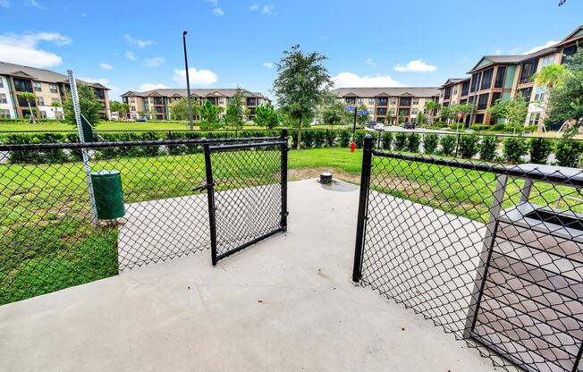 a fenced in dog park with trash cans and apartment buildings in the background