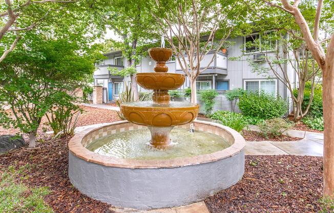 A multi-tiered fountain surrounded by lush greenery and colorful landscaping. The fountain features three levels of water basins with flowing water, set in a circular stone enclosure. In the background, there are modern buildings partially obscured by trees and shrubs.