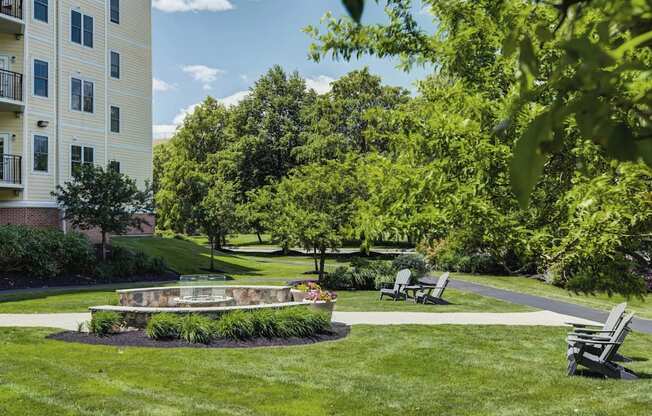 Fire pit with built-in bench seating surrounded by ample green space and trees with part of building in the background.