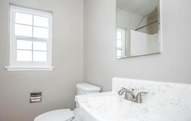 A contemporary bathroom featuring a white marble countertop sink, a mirror, a white toilet, and a window allowing natural light. The walls are painted a soft gray, and there's a glass shower enclosure visible in the background.