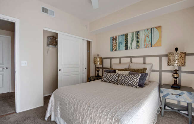 A serene bedroom here at Rio Paradiso featuring a spacious bed dressed in neutral bedding with patterned accent pillows, matching bedside tables with decorative lamps, and soft carpet flooring. The room includes a ceiling fan, an open closet with shelving, and a private doorway leading to an adjoining bathroom for added comfort and convenience.