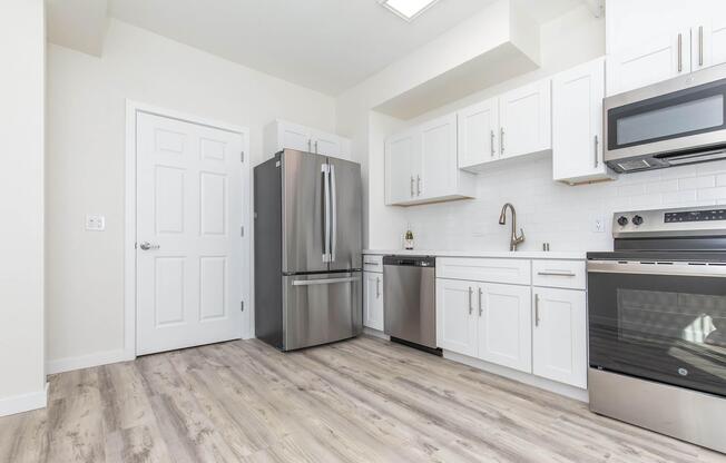 Modern kitchen featuring stainless steel appliances, including a large refrigerator, oven, and dishwasher. White cabinets and countertops complement the light-colored walls and vinyl flooring. A door on the left leads to another room, and there is a window above the sink allowing natural light.