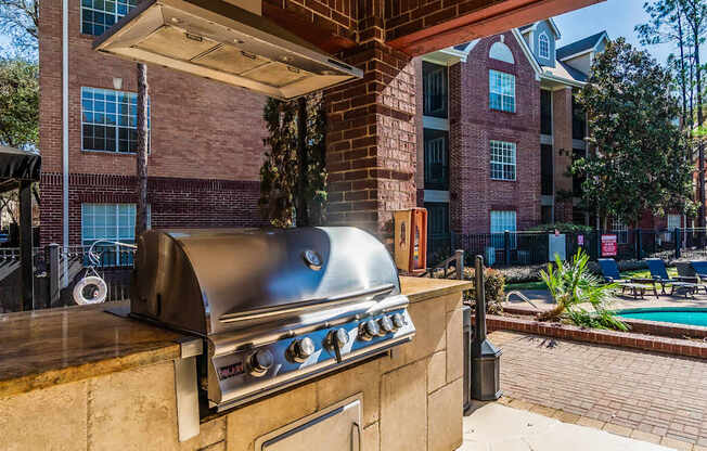 A BBQ grill is on a patio with a pool and trees in the background.